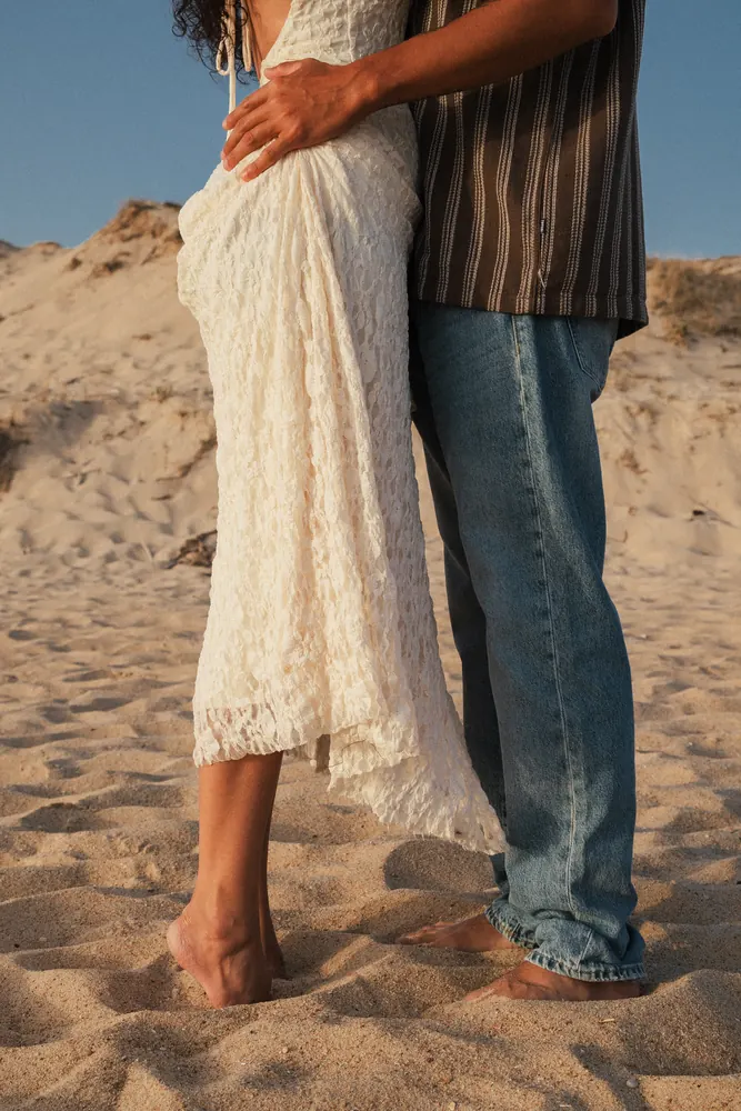 Detalhe de um vestido de renda e pés descalços na areia durante uma sessão de casal